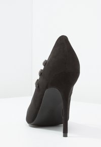 Svart mocka högklackad sko med spetsig tå och tre dekorativa knappar på sidan, med en smal stilettklack.