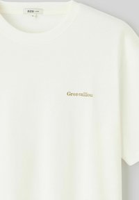 T-shirt blanc à coupe ample en coton, avec un petit texte doré brodé "Gros caillou" sur le côté gauche de la poitrine. Design simple et épuré.