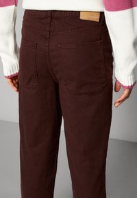 Persona vestida con pantalones marrones con bolsillos tipo parche en la parte trasera y un suéter blanco con mangas a rayas en rosa y morado.