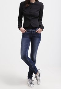 Svart figurnära kavaj med rund hals och knappstängning, paired med slitna blå skinny jeans och metalliska silver sneakers.