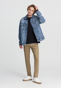 Jasnoniebieska kurtka dżinsowa o klasycznym kroju, z dwiema kieszeniami na piersi, noszona na czarnej koszuli, w zestawieniu z beżowymi spodniami i białymi trampkami.