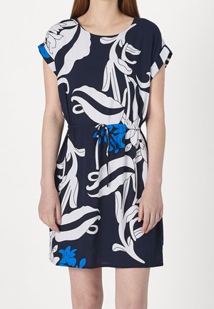Femme portant une robe noire longueur genoux avec des motifs floraux abstraits blancs et bleus et une taille à cordon.