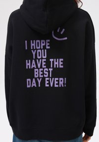 Sudadera negra con un gran texto gráfico en color púrpura en la parte trasera que dice: "¡ESPERO QUE TENGAS EL MEJOR DÍA DE TU VIDA!" y un ícono de cara sonriente.