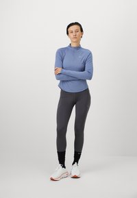 Lichblauwe langarmpul met een halve ritssluiting, gecombineerd met donkergrijze leggings en witte sportschoenen met oranje accenten. Eenvoudig ontwerp en pasvorm.