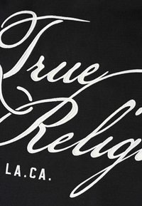 Tissu noir avec texte brodé blanc représentant "True Religion" et "L.A.CA." Le design présente des lettres courbées et des épaisseurs variées.