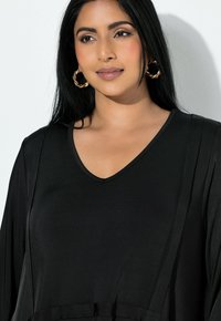 Nainen, jolla on pitkä tummat hiukset, päässään kiertyneet kultarenkki-korvakorut ja musta V-kauluksinen paita, katsoo hieman sivulle valkoista taustaa vasten.