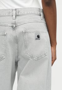 Άτομο που φοράει ανοιχτό γκρι τζιν από denim με ένα μπάλωμα λογότυπο Carhartt στην δεξιά πίσω τσέπη, ορατό από τη μέση μέχρι το μέσο του μηρού.