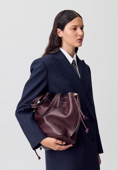 Donna con lunghi capelli scuri, che indossa un blazer blu a righe e una cravatta a righe, tiene una grande borsa a secchiello in pelle bordeaux contro uno sfondo semplice.