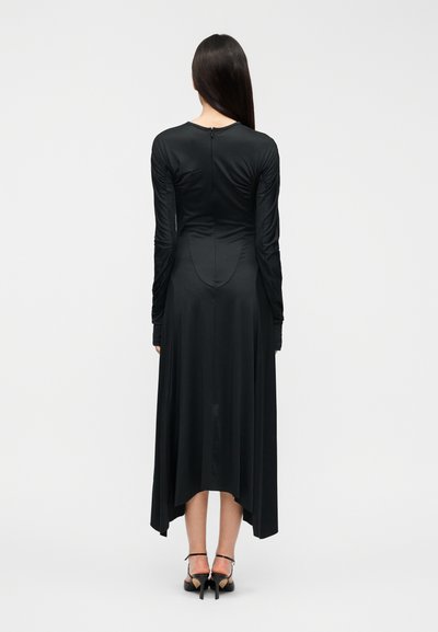Μαύρο φόρεμα μακρυμάνικο με ασύμμετρο τελείωμα, λείο ύφασμα και φερμουάρ στην πλάτη. Συνδυασμένο με μαύρα πέδιλα με λουράκια.