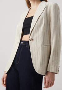 Beige pinstriped blazer with a single button closure, structured lapels, and long sleeves, paired with dark denim high-waisted jeans.