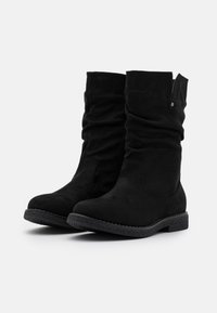 Un par de botas de ante negras de media caña con suelas de goma texturizada de bajo perfil y detalle de caña ligeramente arrugada, sobre un fondo blanco liso.