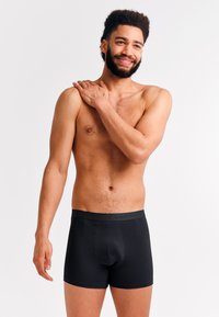 Bearded man with curly hair wearing black boxer briefs stands against white background, smiling and touching his left shoulder.
