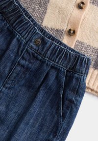 Dunkelblaue Jeanshose mit elastischem Bund, kombiniert mit einer beigefarbenen und grauen gemusterten Strickjacke mit braunen Knöpfen.