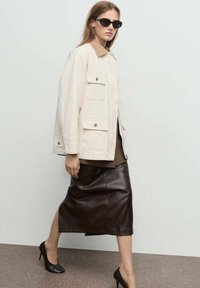 Crème-kleurige, oversized jas met een beige kraag, gedragen over een bruine top en een bruine leren kokerrok, gecombineerd met zwarte hoge hakken.