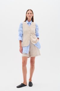Bežs lina veste virs gaiši zila krekla ar pogu akcentiem, kombinēta ar gaišiem šortiem un melniem slip-on apaviem. Vienkāršs, pielāgots dizains.