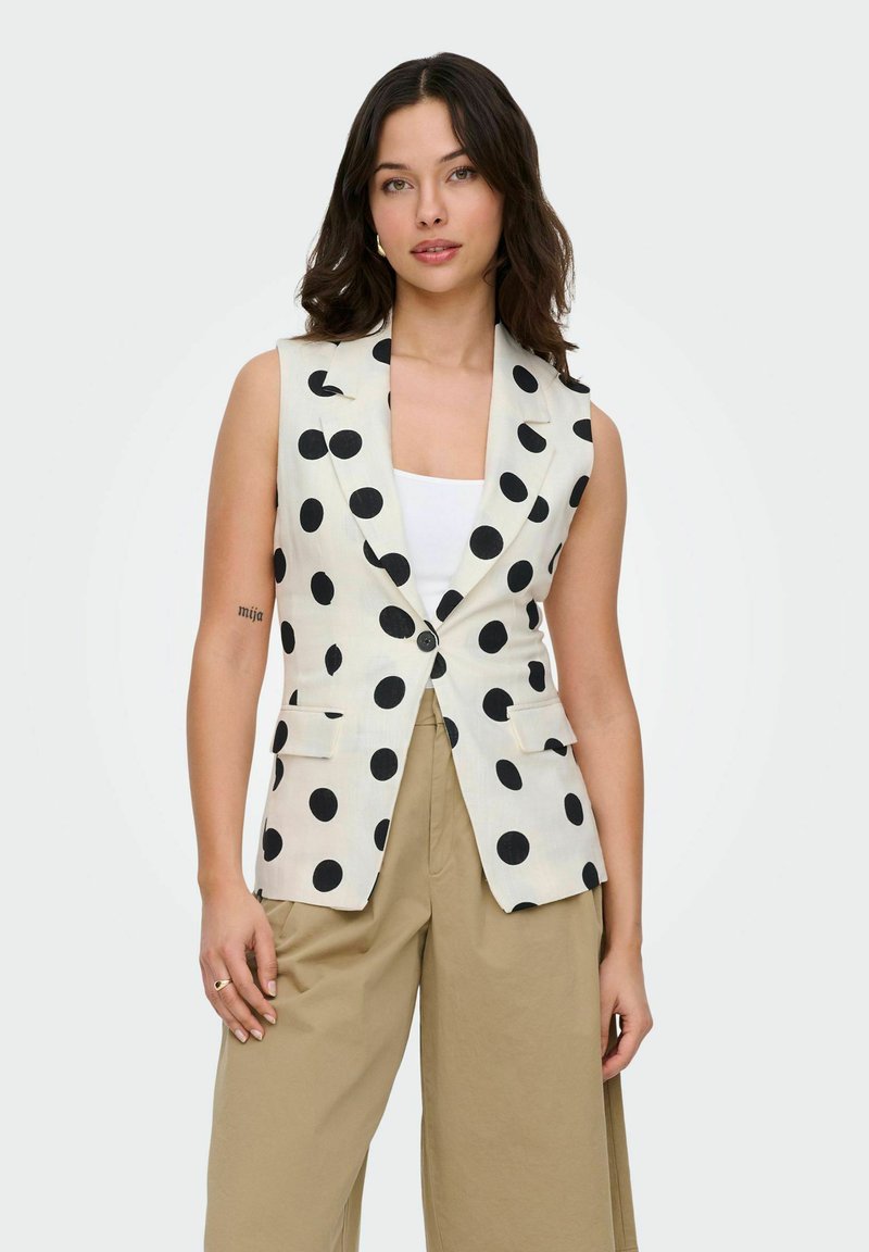 Femme aux cheveux foncés portant un blazer blanc sans manches à gros pois noirs, un haut blanc et un pantalon beige ample, debout, les mains détendues.