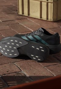 Czarne buty sportowe z teczowymi paskami, teksturowana cholewka oraz wzorzyste gumowe podeszwy z białymi okrągłymi akcentami. Ustawione na ceglastej powierzchni.