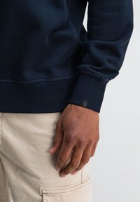 Marineblauwe sweatshirt met ribbelboorden en onderrand, met een gestikte textuur en een klein zwart etiket op de mouw. Lichtbeige broek zichtbaar.