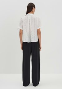 Blusa bianca a maniche corte con linee verticali e una piega sul retro, abbinata a pantaloni neri a gamba larga, su uno sfondo neutro.