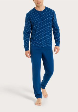 Uomo che cammina a piedi nudi indossando una maglia blu a maniche lunghe con puntini bianchi e pantaloni blu tinta unita abbinati su sfondo neutro.