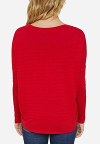Rød langærmet sweater med ribbet tekstur, bådudskæring og en afslappet pasform. Har sideslids og en lidt længere bagkant.