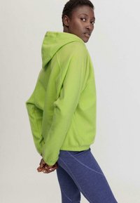 Grüne Kapuzenjacke aus weichem Fleece; verfügt über einen lässigen Schnitt und eine Kängurutasche, kombiniert mit blauen Leggings mit kontrastierenden Nähten.