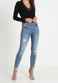 Schwarzer langärmeliger Bodysuit mit Spitze am Ausschnitt und an den Handgelenken, kombiniert mit hellblauen, zerstörten Skinny-Jeans und blauen Absatzsandalen.