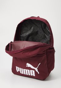 Burgundin väri selkäreppu, jossa on iso etutasku, jossa on Puma-logo. Silkkinen kangaspinta ja musta kuviollinen vuori sisällä.