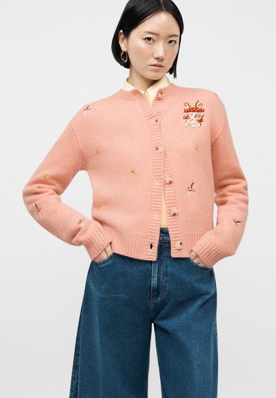 Γυναίκα που φοράει ένα ροζ ζακέτα με κουμπιά, διακοσμημένο με μικρά κεντητά μανιτάρια και λουλούδια, συνδυασμένο με φαρδιά μπλε τζιν παντελόνια.