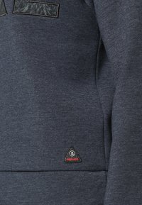 Primo piano di una felpa grigio scuro con un piccolo patch triangolare "FIRE+ICE" vicino all'orlo sul lato anteriore.