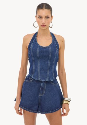 Femme portant un haut sans manches en denim à col halter et un short en denim taille haute, accessoirisée avec de grandes boucles d’oreilles créoles et des bracelets en or.