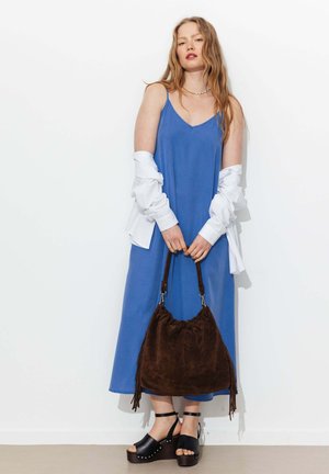 Femme en robe nuisette bleue avec une chemise blanche tombant des épaules, tenant un grand sac à main en daim marron, portant des sandales à plateforme noires.
