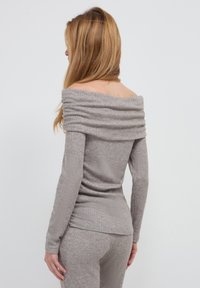 Frau mit langen Haaren, die ein graues, geripptes Off-Shoulder-Oberteil mit langen Ärmeln und dazu passende gerippte Hose trägt, mit dem Rücken zur Kamera vor einem einfarbigen Hintergrund.