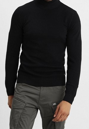 Homme portant un pull noir structuré ajusté et un pantalon cargo gris avec des poches zippées et à rabat, debout avec une main près de la poche.
