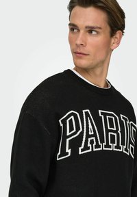 Pull noir avec un grand texte blanc "PARIS", col rond et motif en maille texturée sur les épaules.