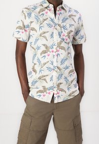 Camisa de manga corta abotonada, confeccionada en tela ligera y con un estampado floral tropical en tonos rosa, verde y marrón. Incluye un bolsillo en el pecho.