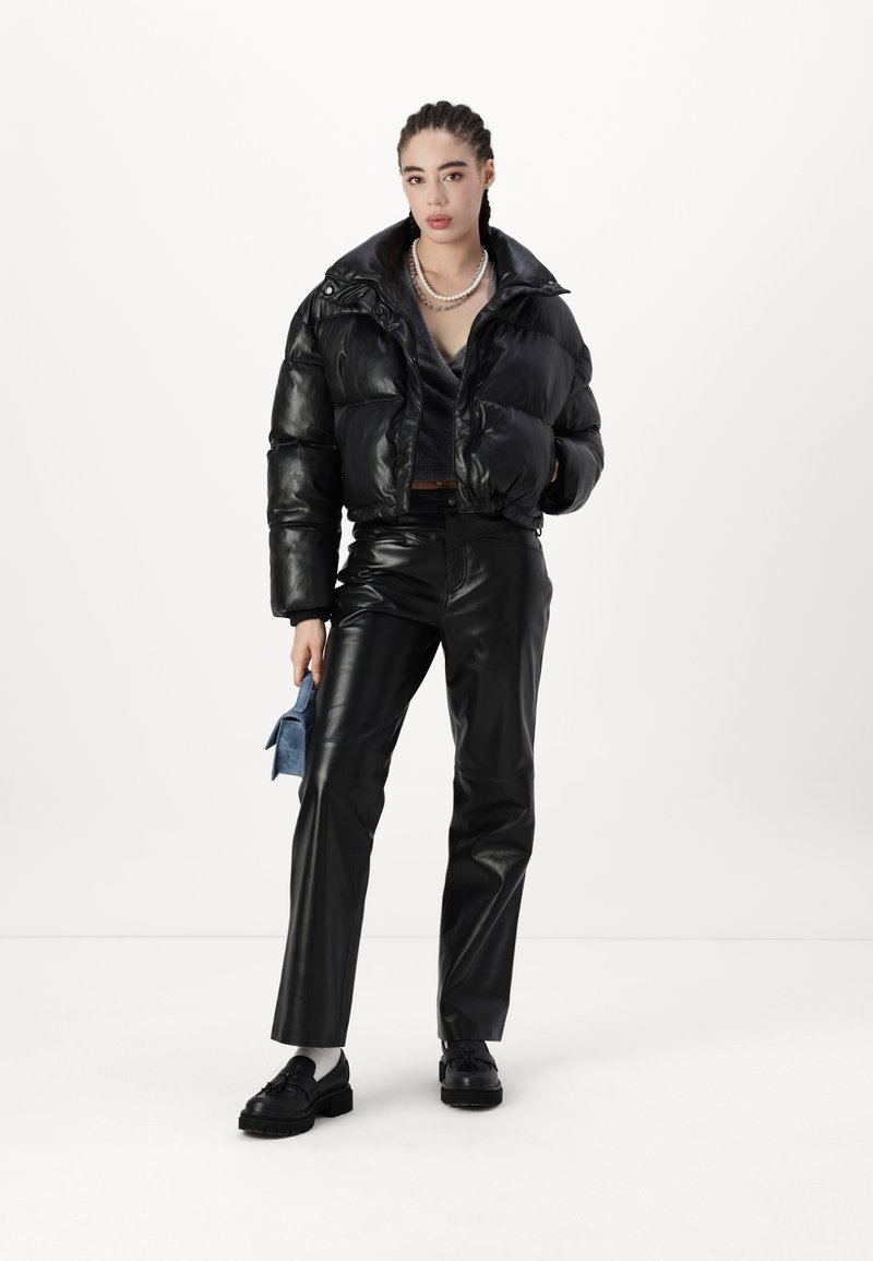 Schwarze Pufferjacke mit glänzender Oberfläche, taillierte Form und hohem Kragen. Kombiniert mit schwarzen Kunstlederhosen und schwarzen Schuhen.