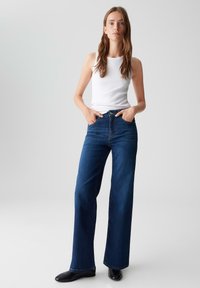 Giovane donna in piedi con le mani nelle tasche, che indossa una maglietta bianca senza maniche, jeans a gamba larga blu scuro e scarpe nere, su uno sfondo neutro.