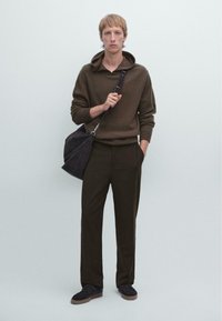 Bruine gebreide hoodie met een relaxte pasvorm, donkere broeken en zwarte suède sneakers. Het model houdt een zwarte stoffen schoudertas vast.