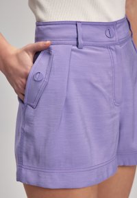 Shorts lilas à taille haute, avec poches latérales et détails en boutons. Texture lisse et coupe ajustée avec des coutures contrastées.