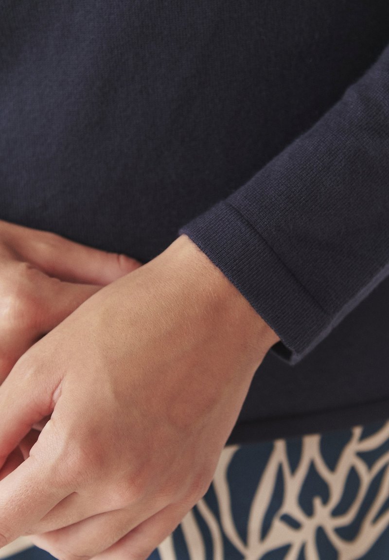 Granatowa koszula z długim rękawem z teksturowanej tkaniny i dopasowanym mankietem, w połączeniu ze wzorzystą odzieżą w niebiesko-białe wzory. Ręce na biodrach.