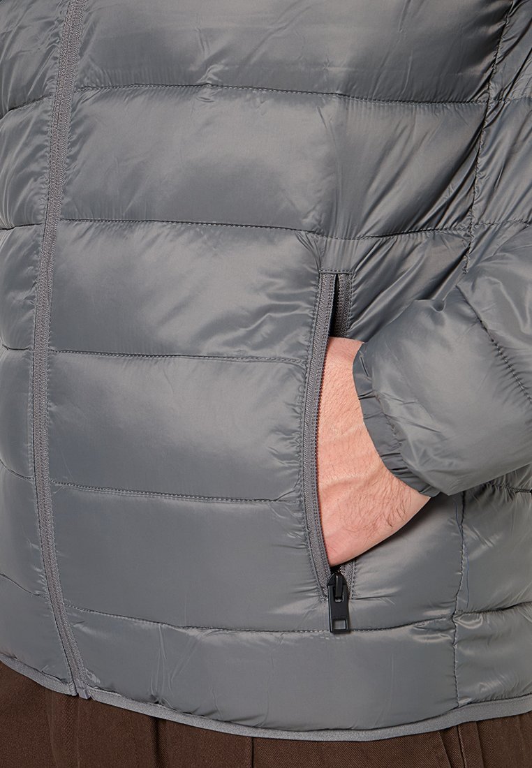 Chaqueta acolchada gris hecha de material brillante y ligero. Presenta un bolsillo con cremallera y puños elásticos. La costura resalta el diseño acolchado.