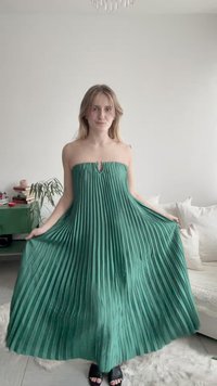 Mujer de pie en interiores vistiendo un vestido verde plisado sin tirantes, sosteniendo los lados, con una planta, libros y un sofá blanco en el fondo.