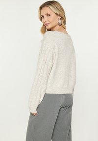 Lichtbeige trui met een zachte, gestructureerde afwerking, voorzien van geribbelde kabelgebreide mouwen en een gegolfde halslijn. Gecombineerd met grijze wijde broek.