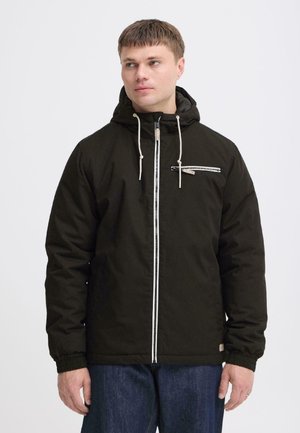 Man wearing a black hooded jacket with a white zipper and zippered chest pocket, looking to his left against a plain background.