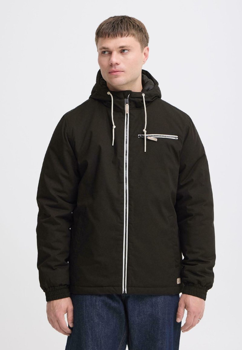 Homme portant une veste à capuche noire avec une fermeture éclair blanche et une poche poitrine zippée, regardant vers sa gauche sur un fond uni.