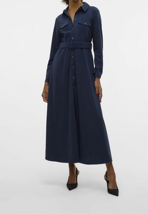 Frau trägt ein langes, marineblaues Hemdblusenkleid mit Gürtel und Knopfdetails sowie schwarze spitze Slingback-Pumps vor einem einfarbigen Hintergrund.