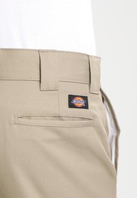 Beige kalhoty vyrobené z pevné látky, s černou značkou Dickies na zadní kapse a tradičním designem pásku.