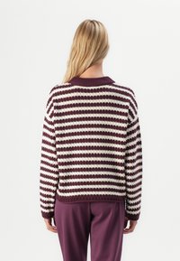 Blond inimene seljaga, kandes tekstuuriga lilla ja valge horisontaalselt triibulist sviitrit ning ühtlasi lillat pükse lihtsa tausta ees.