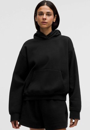 Frau mit dunklen Haaren trägt einen schlichten schwarzen übergroßen Hoodie und passende schwarze Shorts vor einem hellgrauen Hintergrund.